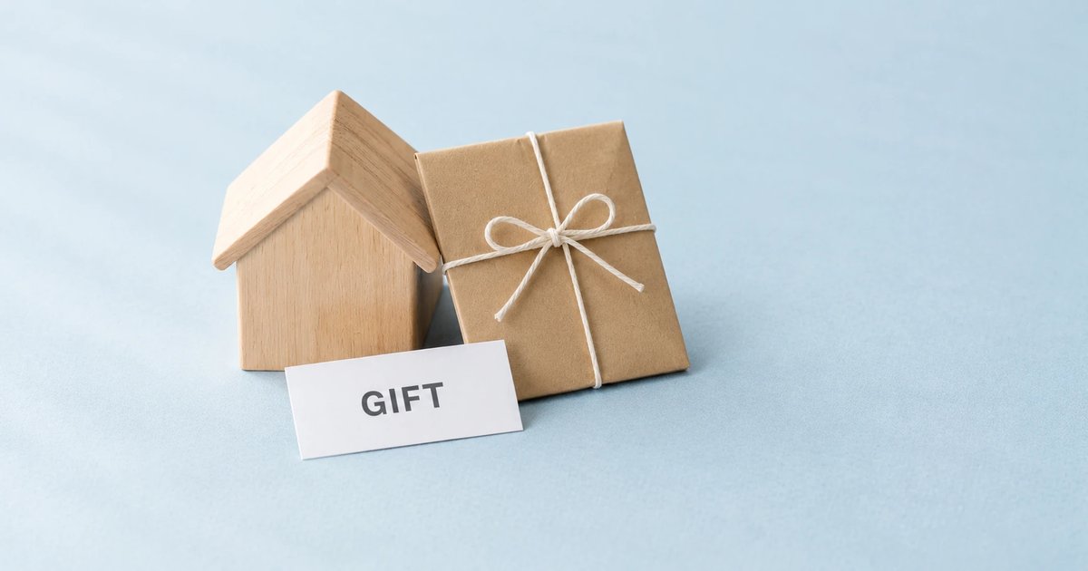 淡い空色の紙の上に、木の家の積み木と、ひもで結んだクラフト紙の包み。手前に「GIFT」のカード